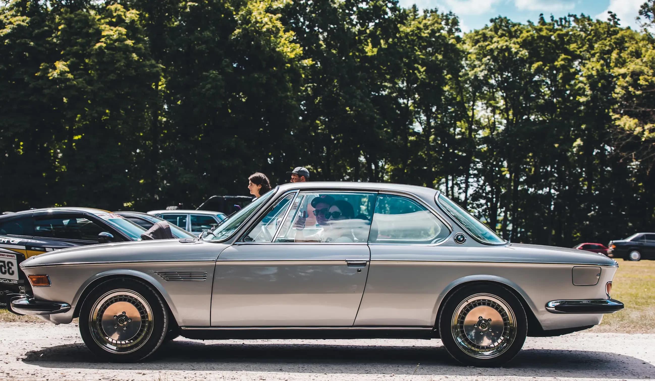 A silver classic car parked outdoors with green trees in the background
