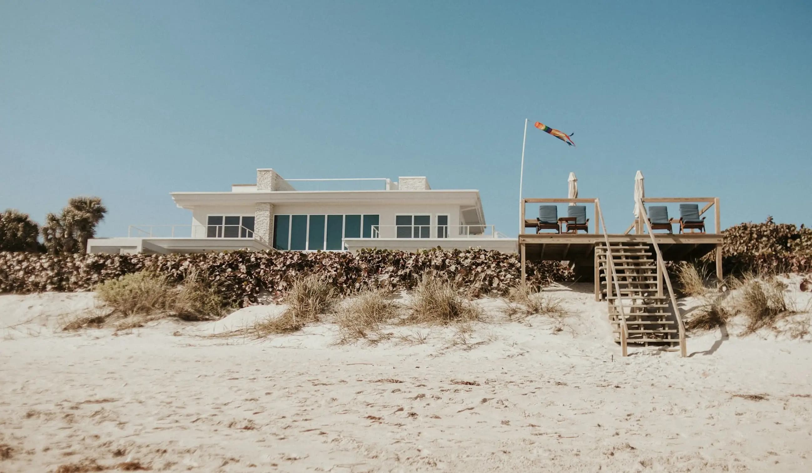 Modern beach house with large glass windows and a wooden deck, surrounded by sand and coastal vegetation
